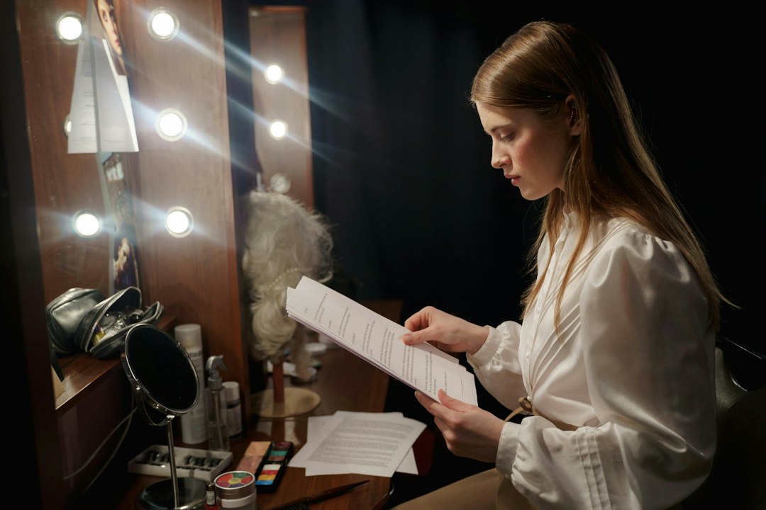 A woman in a white blouse studies a stack of printed pages at a dressing-room table lit by bulb lights, rehearsing before a performance.