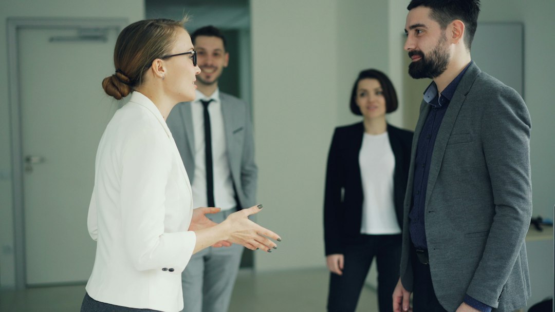 A woman in a white blazer gestures with open hands as she speaks to a bearded colleague in an office while two coworkers look on