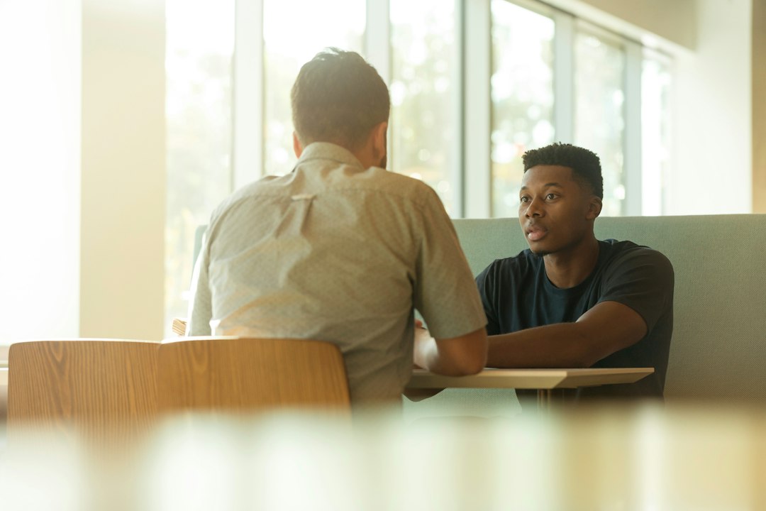 A young man in a dark t-shirt speaks across a small table to an interviewer seen from behind, in a bright room with tall windows.