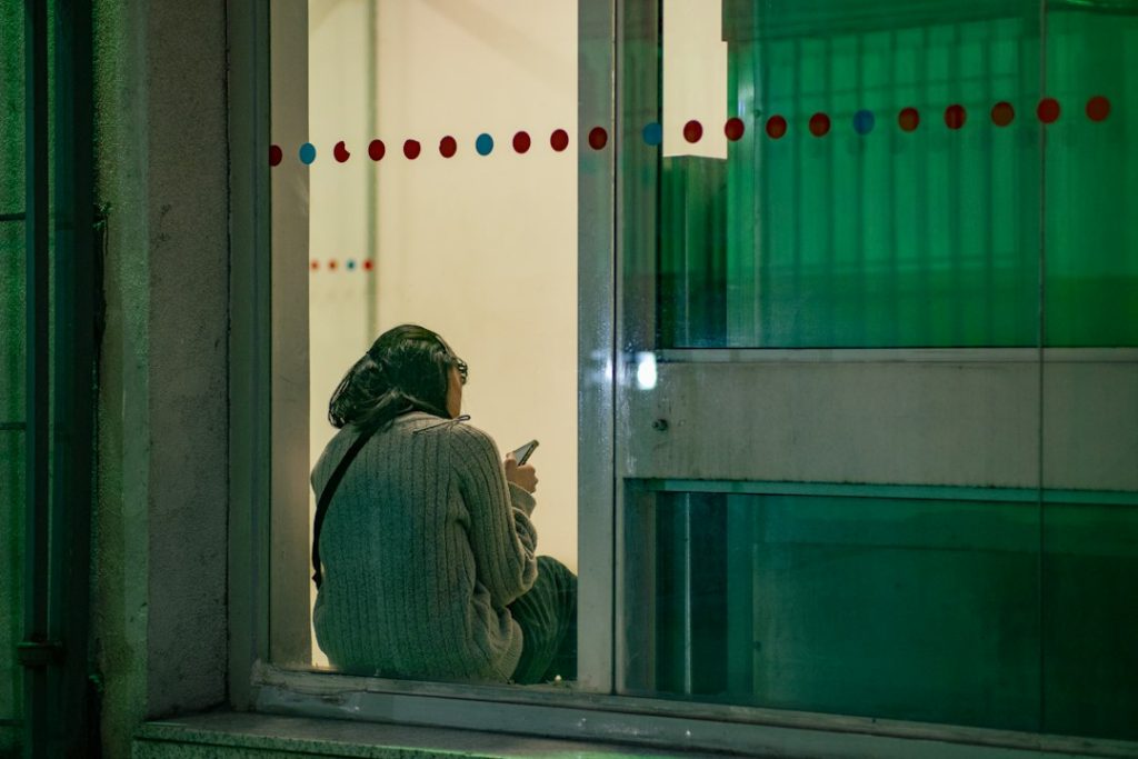 A woman sits on a window ledge at night, turned away, looking at her phone as she waits for a message