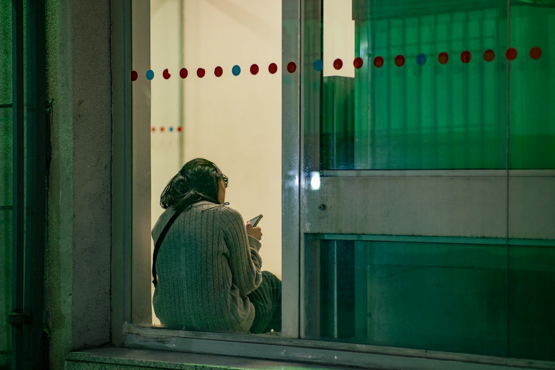 A woman sits on a window ledge at night, turned away, looking at her phone as she waits for a message