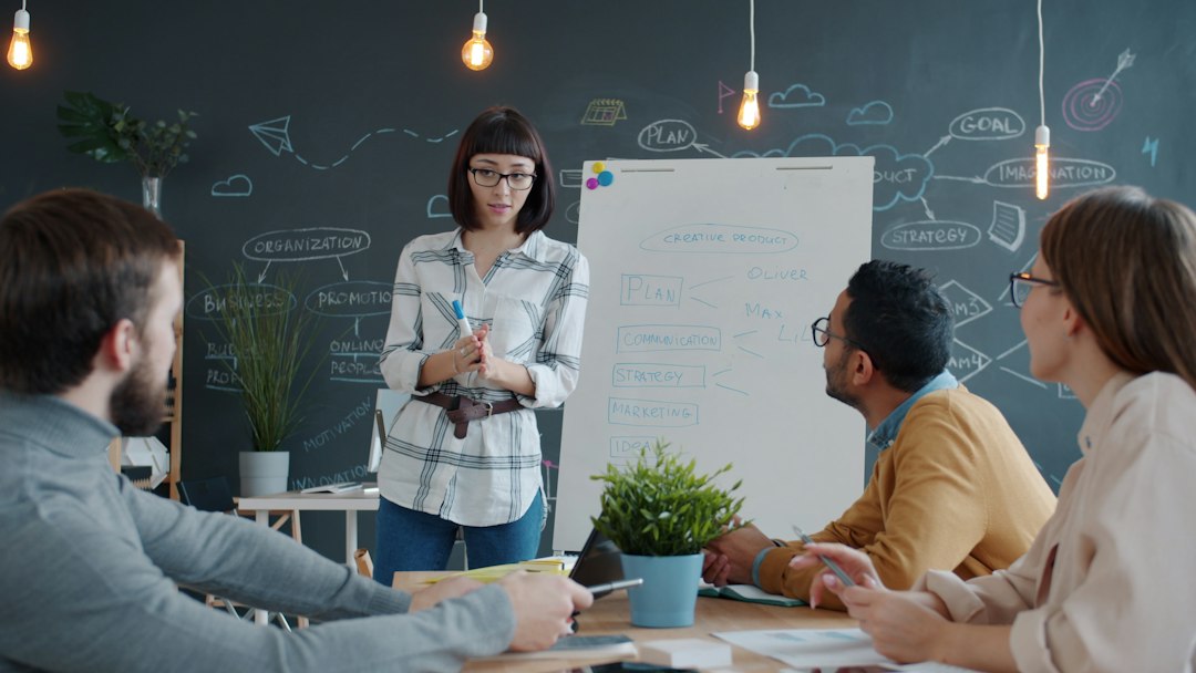 A woman in glasses stands beside a whiteboard reading Plan, Communication, Strategy and Marketing, explaining her thinking to three colleagues seated at a table.