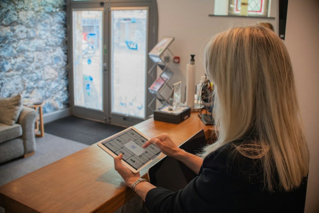A woman seated at an office reception desk, holding a tablet beside the front door and waiting area