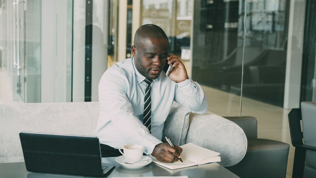 A man in a light shirt and striped tie takes a phone call while writing notes in a notebook, with a laptop and coffee cup on the table in front of him