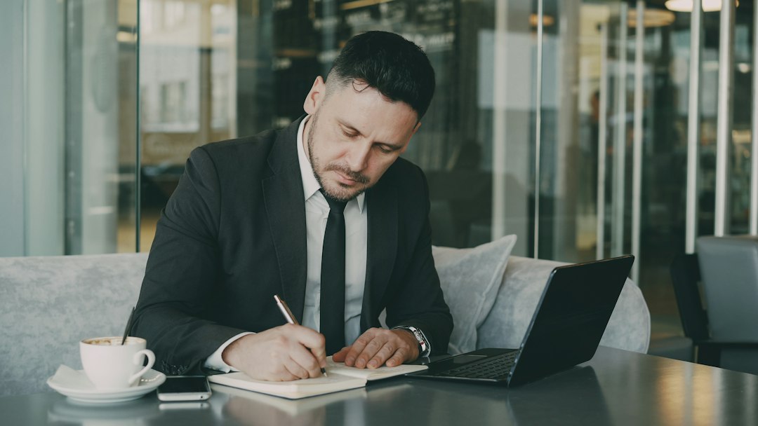 A man in a dark suit writing in a notebook at a cafe table, a laptop and coffee beside him, the way an interviewer takes notes on a candidate