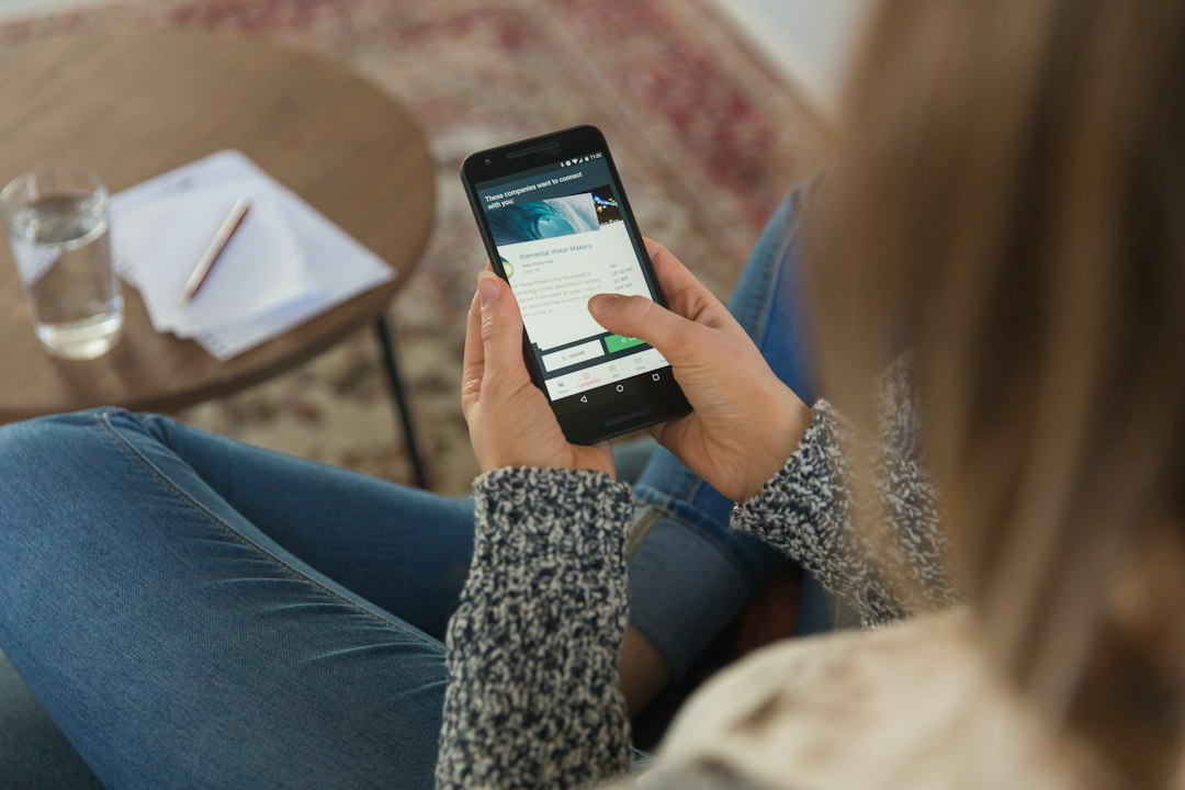 Job seeker sitting on a rug scrolling a phone app that lists companies hiring, with a notebook and glass of water on a side table